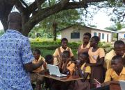 A librarian teaching a group of school children to learn computer skills. The class is taking place outside, under a tree.