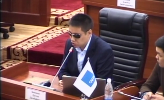 MP Dastan Bekeshev at a desk, addressing the meeting, through a microphone