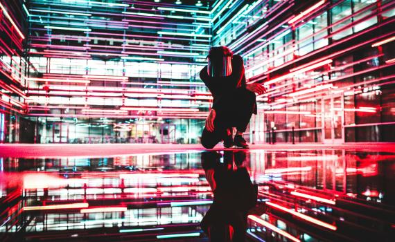Image by Drew Dizzy Graham, Unsplash, https://unsplash.com/license Image of a man kneeling inside a building, with neon lights and reflections, to suggest many dimensions of AI. Image by Drew Dizzy Graham, Unsplash, https://unsplash.com/license