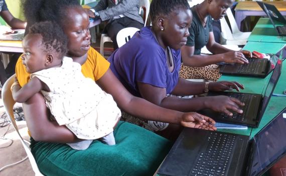 A young woman learning computer skills, with a baby on her shoulder.