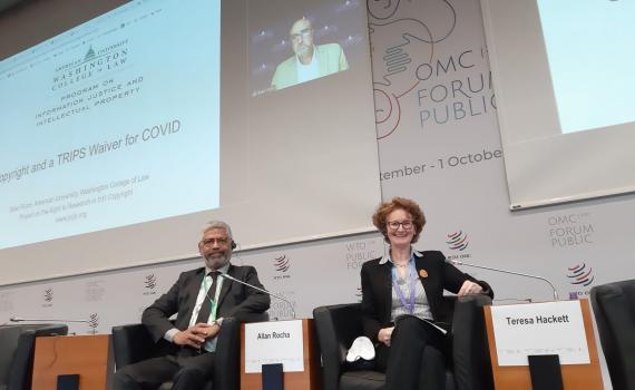 Allan Rocha and Teresa Hackett participating in a working session at the WTO Public Forum. They are seated, in front of a large screen where information / visuals are projected.